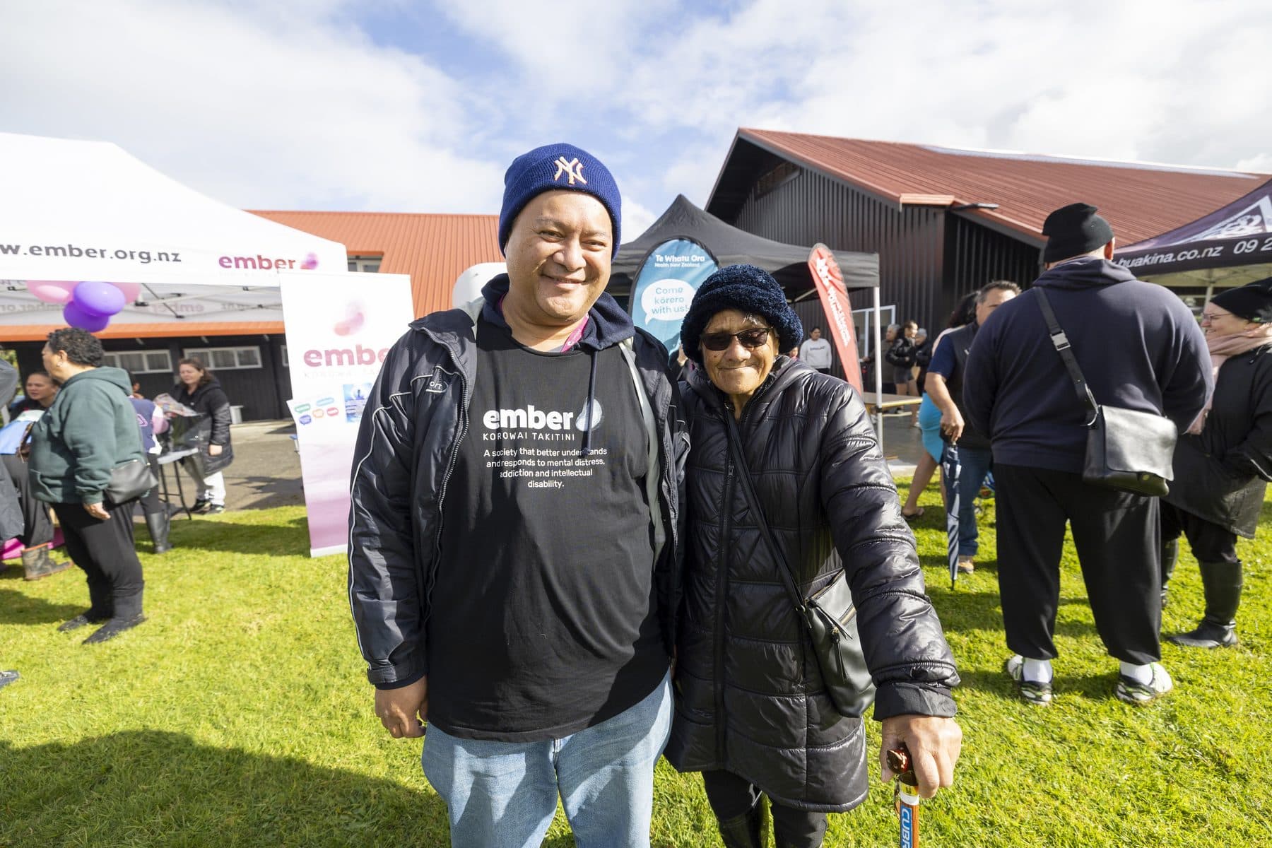 A man and an older woman standing together and smiling on the grass, with marquees behind them.