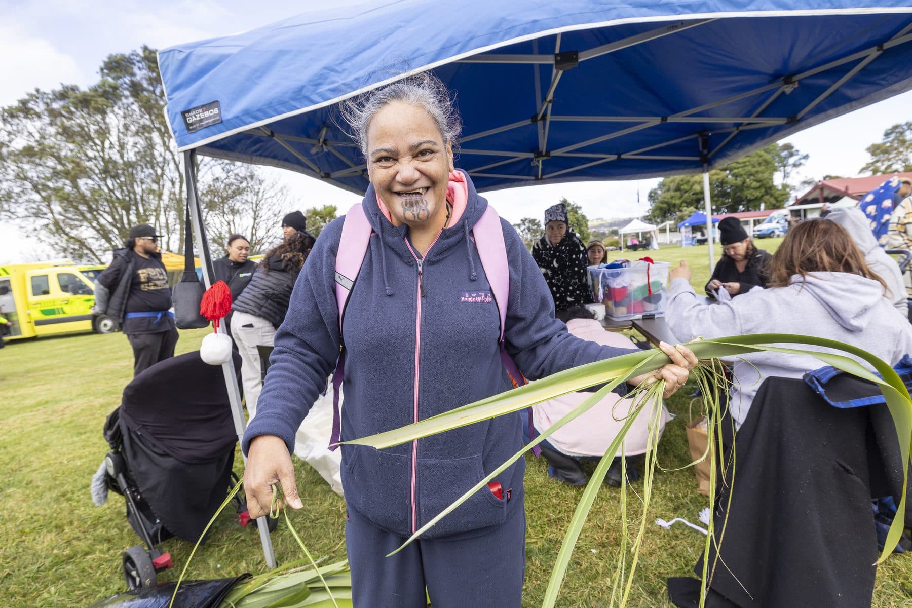 A woman smiling and holding strips of harakeke under a gazebo at a community day.